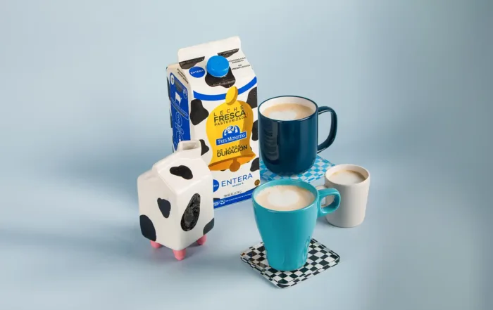 Bote de Leche Tres Monjitas de larga duración entera con tecnología MST, rodeada de tazas con diferentes diseños.