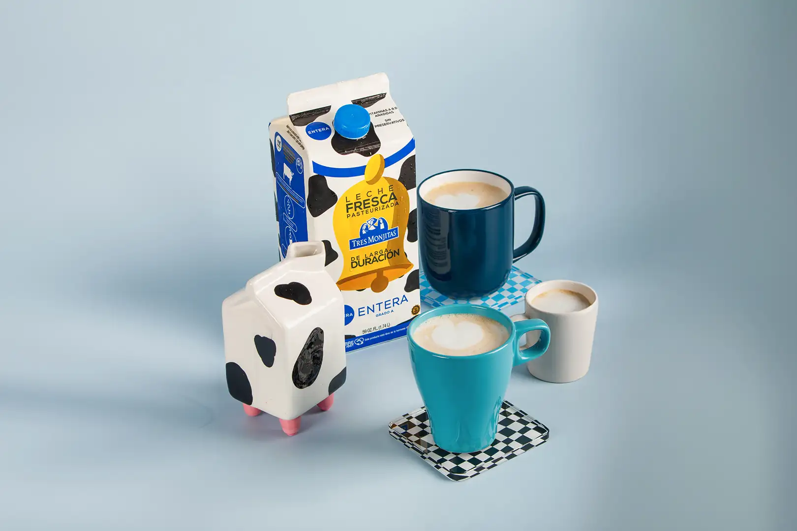 Bote de Leche Tres Monjitas de larga duración entera con tecnología MST, rodeada de tazas con diferentes diseños.